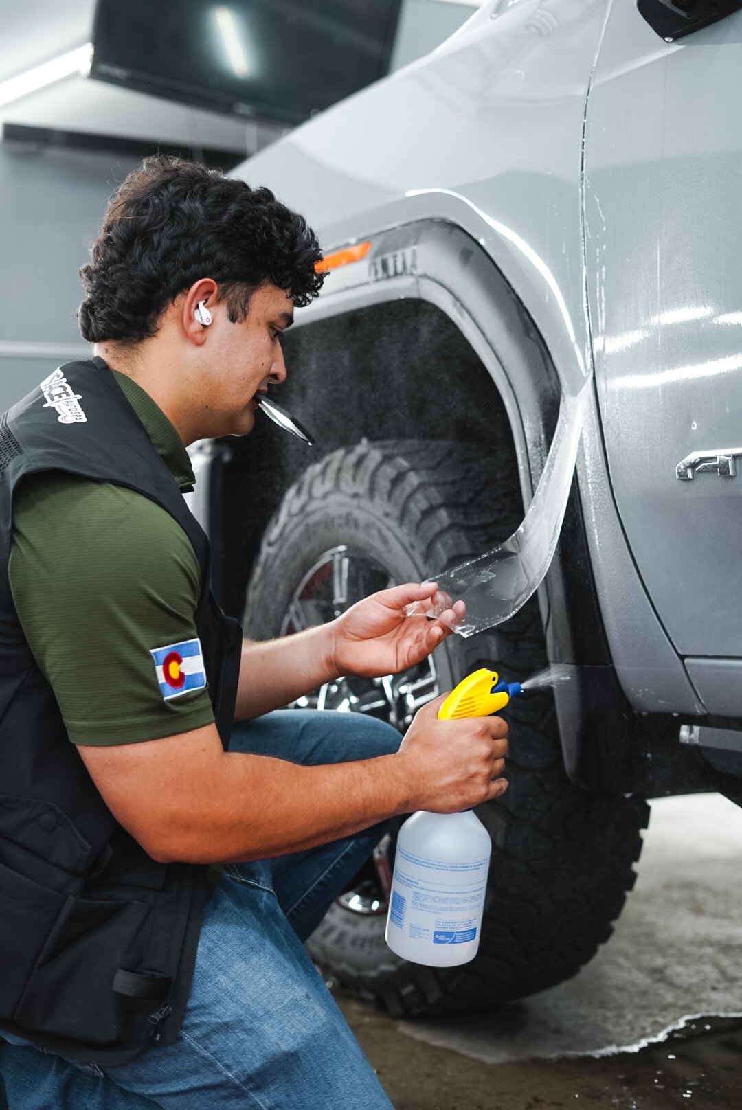 Technician applying paint protection film to the wheel arch of a silver truck, Colorado flag patch visible on jacket